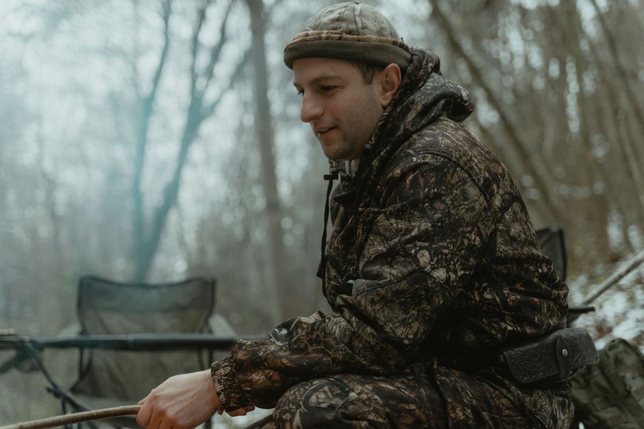 A man in camouflage gear sits in a serene forest, blending seamlessly with nature.
