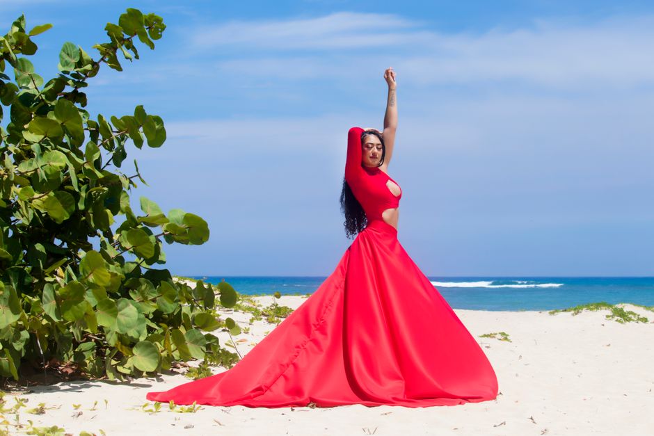 Fashion photoshoot of a woman in a red dress posing elegantly on a sunny Caribbean beach.