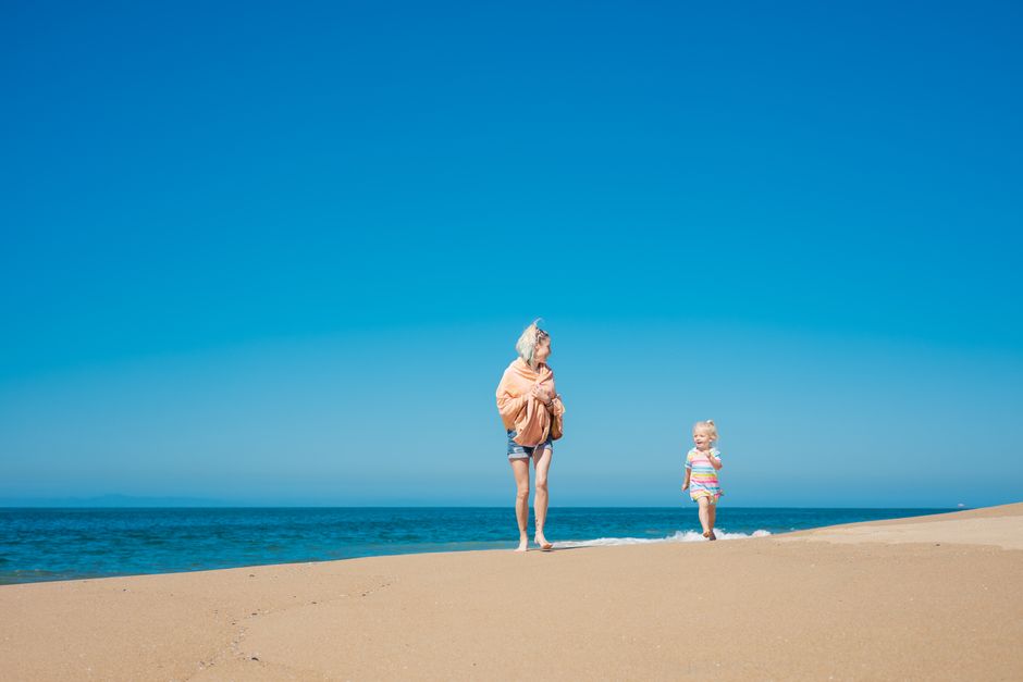 A mother and daughter walking on a sunny beach with a clear blue sky.