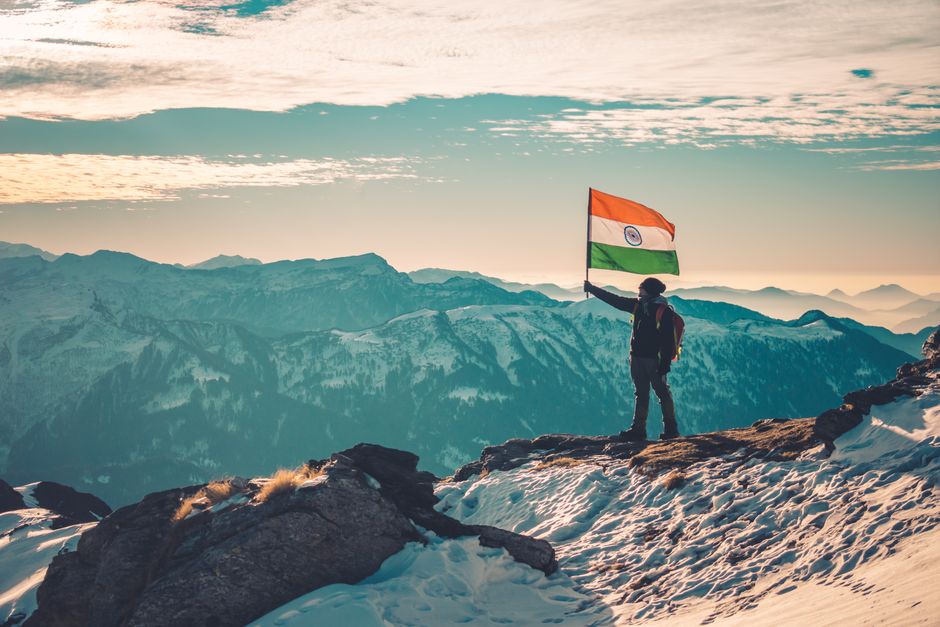 Man holding Indian flag atop a scenic snowy mountain range, capturing nature's beauty.
