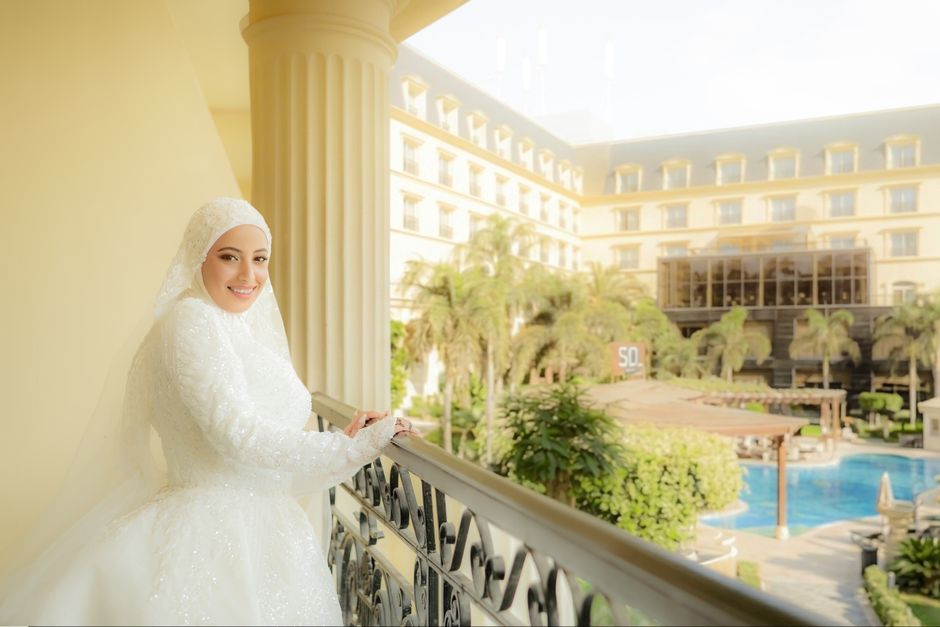 A radiant bride in a white dress stands on a balcony overlooking a luxurious hotel pool area.