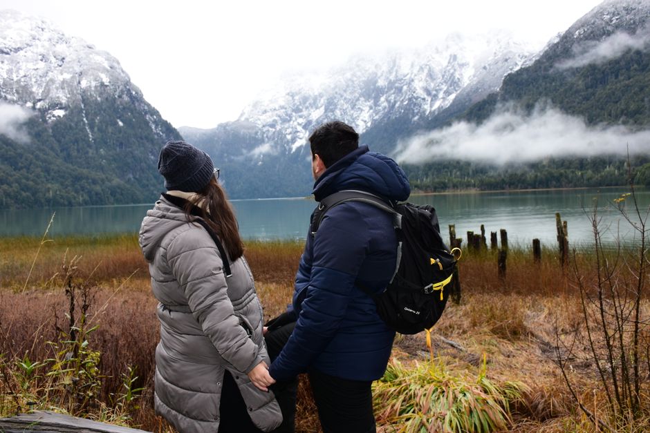 A couple in winter clothing enjoying a tranquil view of a mountain lake surrounded by snow-capped peaks.