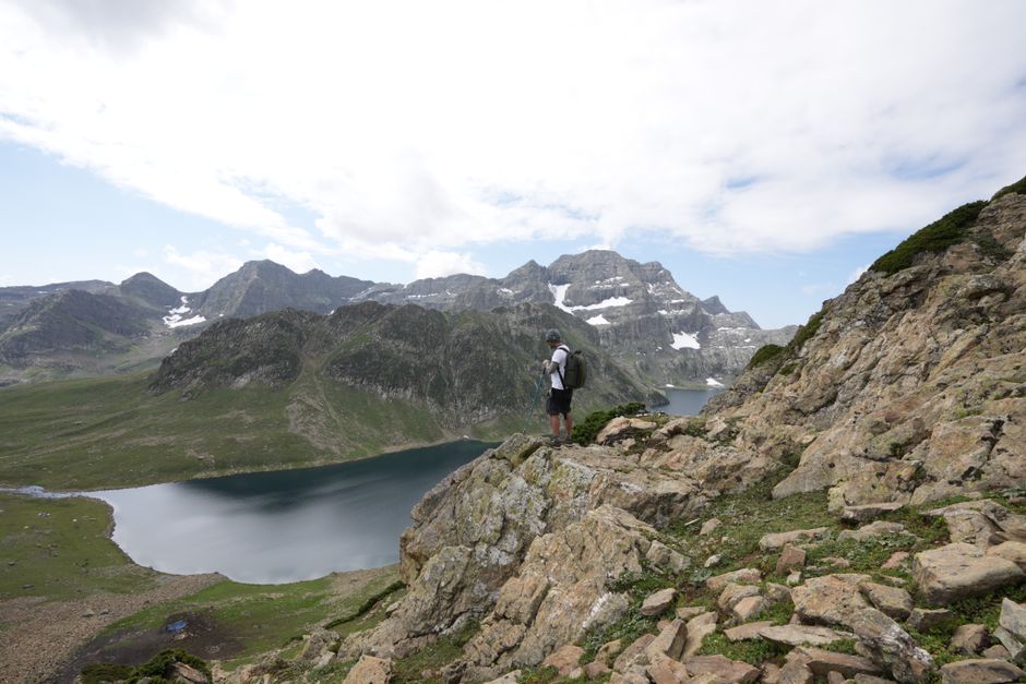 A hiker stands on a rocky cliff overlooking a serene mountain lake under a partly cloudy sky.