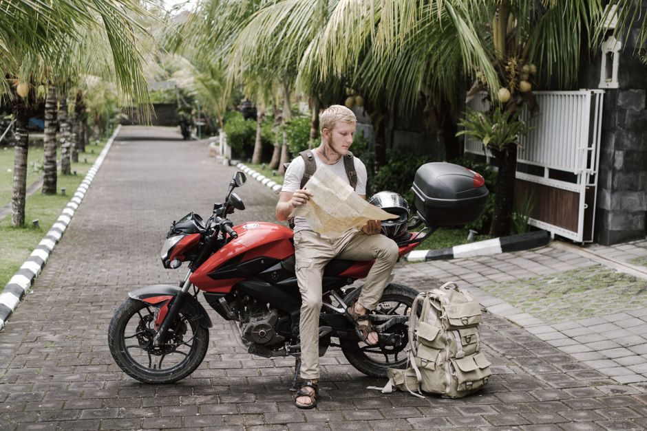 Full body male tourist in casual clothes with backpack helmet and duffle bag studying city map while leaning on parked motorbike on street under palms