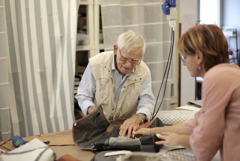 Concentrated elderly craftsman in casual clothes standing at table in workshop and making leather accessory with help of female partner