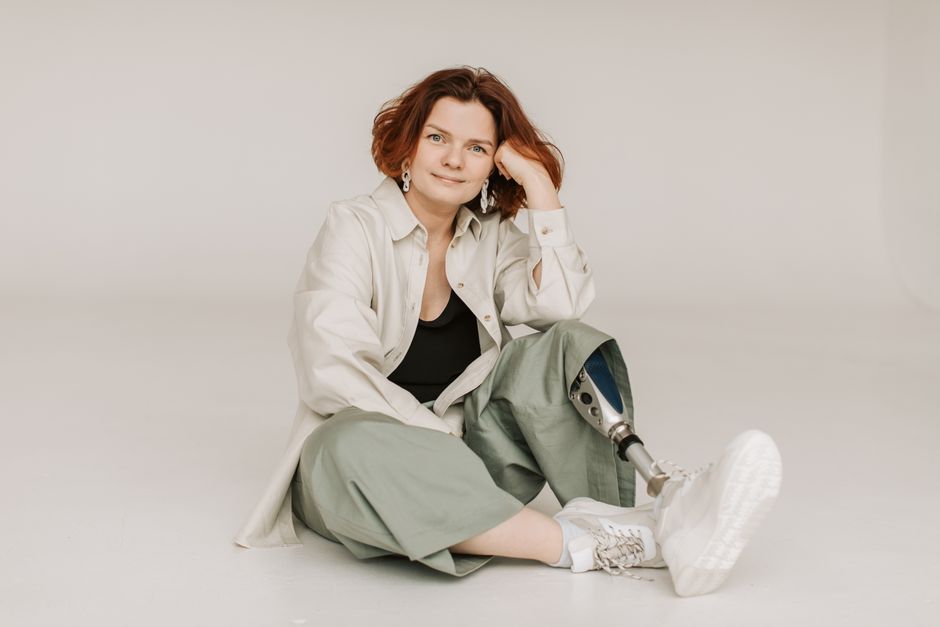 Confident woman with prosthetic leg sitting indoors on a white background.