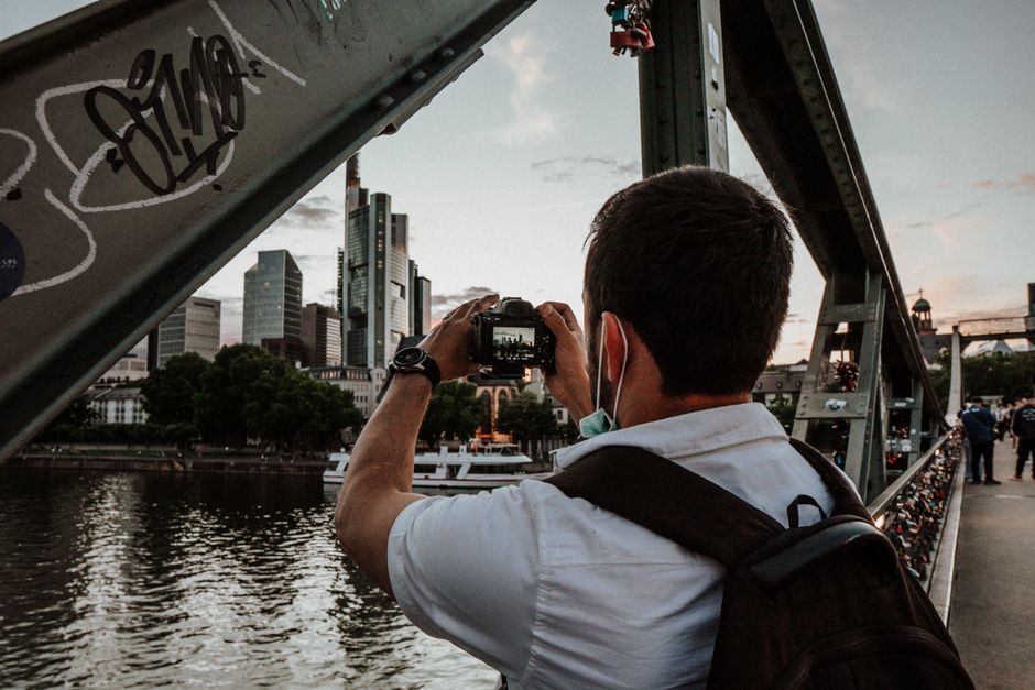 Man on bridge photographing urban skyline at sunset.