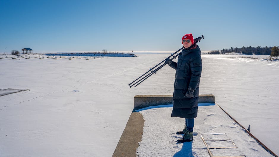 A photographer stands on a snowy pier holding a tripod, capturing winter scenery.