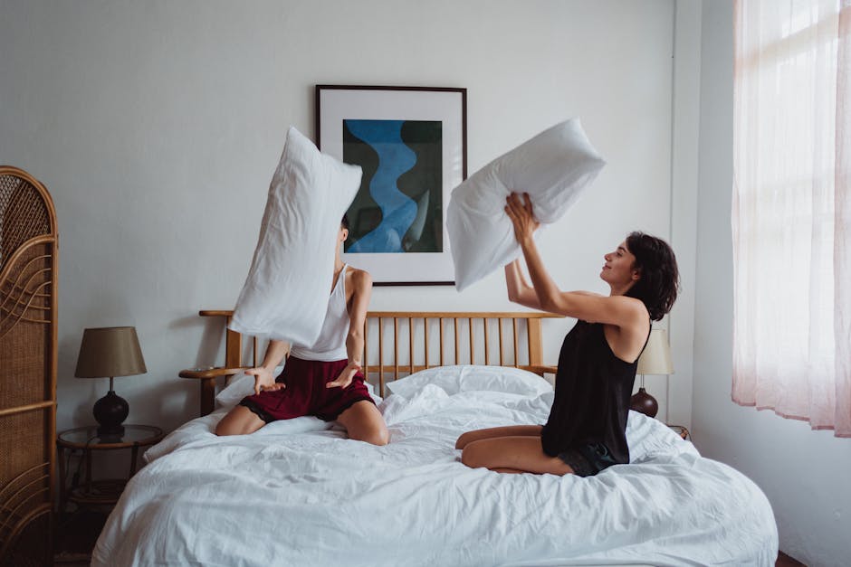 Two women enjoying a playful pillow fight on a bed in a cozy bedroom setting.