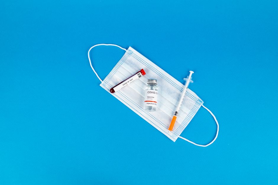 Flat lay of COVID-19 vaccine vial, syringe, and mask on blue background, highlighting pandemic essentials.