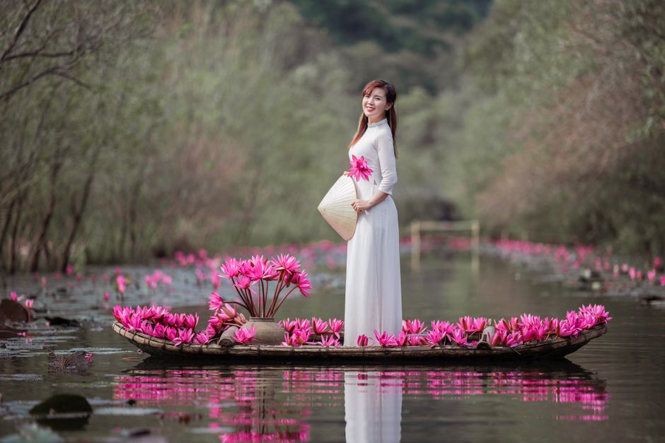 Asian woman standing on a lotus-decorated boat in a tranquil river setting.