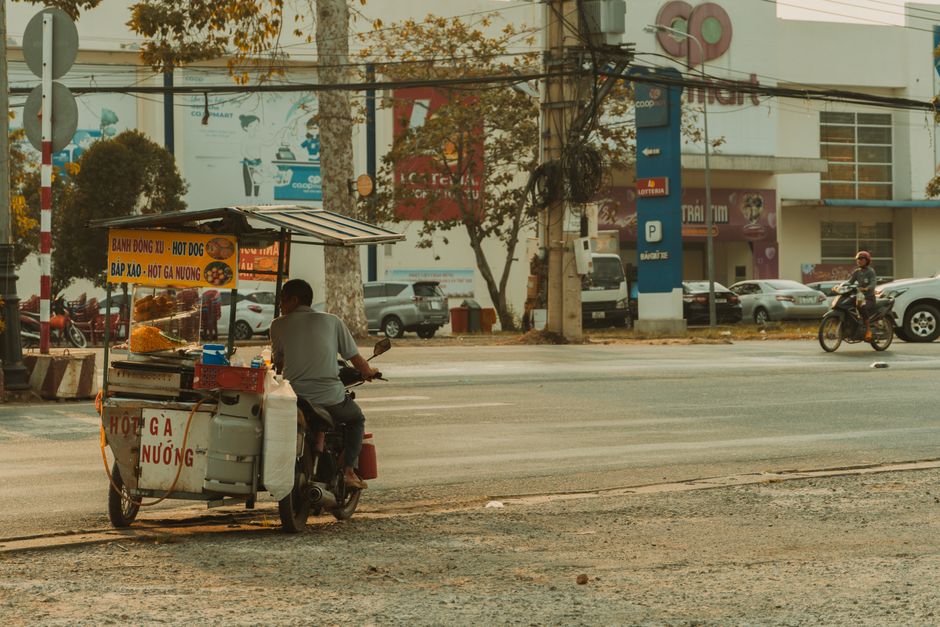 A street food vendor rides a modified motorbike cart selling snacks on a city street corner.